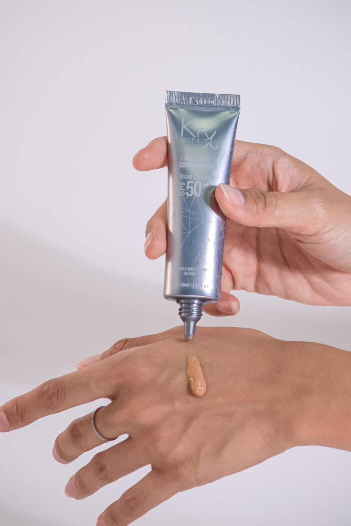 KrX Aesthetics Tinted Sun Protection – eine Hand hält die Tube, während die getönte Creme auf den Handrücken aufgetragen wird, SPF 50+ PA++++, 30 ml.